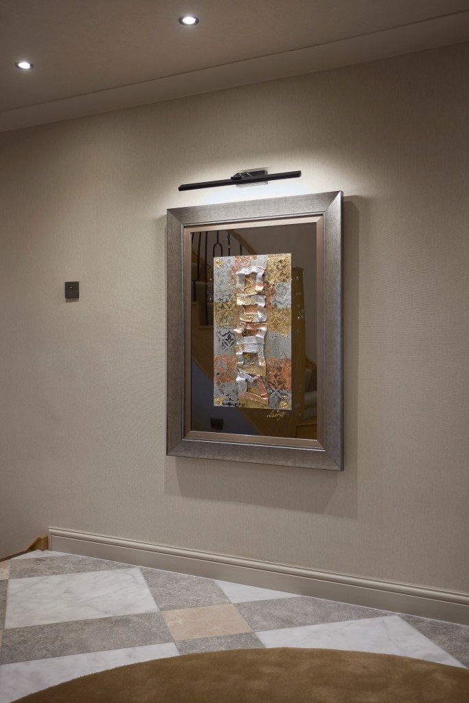 A large silver-framed mixed media artwork with rust and gold tones illuminated by a black picture light above a checkered marble floor.