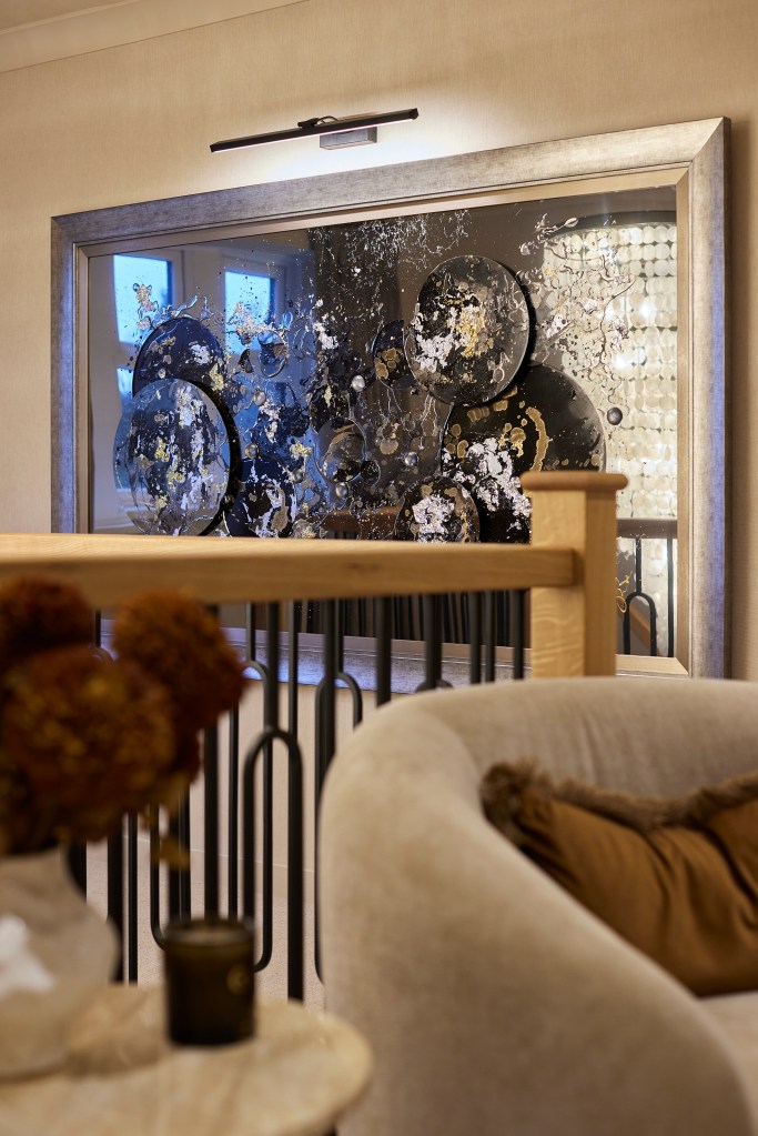 A neutral velvet armchair with a rust cushion in front of a large, silver-framed abstract mixed media artwork.