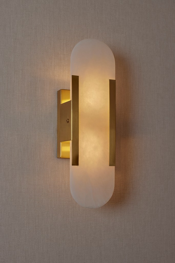 A vertical, modern gold wall sconce with an alabaster-style shade mounted on neutral textured wallpaper.