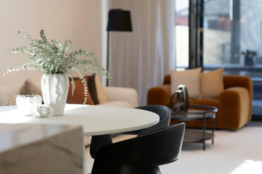 A modern, multi-functional dining zone featuring a small round white table and black chairs, set against a blurred background of a warm, velvet-upholstered living area. The space illustrates open-plan living and the transition from formal dining.